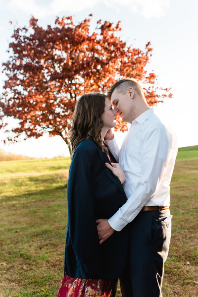 Young couple lean in for a kiss at Mingo Creek County Park in Finleyville, standing in front of a tree whose leave are lit up red from the fall sunset and she wears his suit jacket over her shoulders during their engagement session.