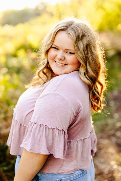A person with long, wavy hair smiles at the camera in a sunlit outdoor setting captured by Amber Bauer Photography. They wear a light purple top with flared sleeves and blue jeans, set against a bright backdrop of greenery.