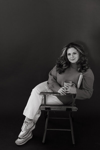 Sara Dobbins sits in a chair facing sideways while smiling at the camera and holding a mug