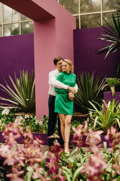 Woman wearing a green dress with fiancé in colorful garden in front of a purple & pink building.