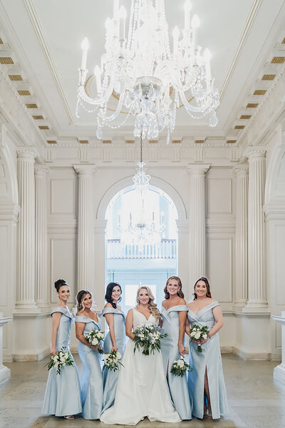 Bride stands with bridesmaids in icy blue gowns under a large chandelier in an elegant cream room with soaring columns.