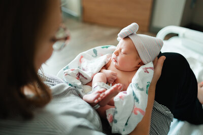 The newborn's mother in the photograph is holding him with both hands. The newborn is wearing a white hat with colorful stripes and is covered with a white blanket.