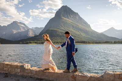 Bride leads Groom along a rock wall next to a lake with giant mountains across the lake in Glacier National Park