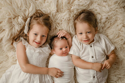 Two older siblings snuggling with their newborn baby sister during a cozy studio family session at Duehr Photography in Maple Grove, Minnesota