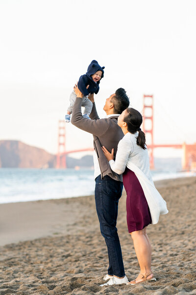 Dad holding up baby in front of the golden gate bridge while mom snuggles up behind him