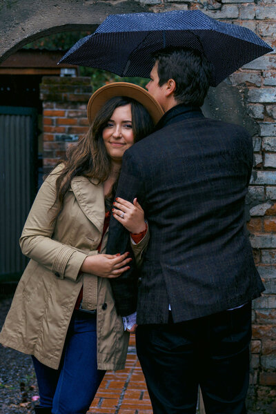Engaged couple sharing a quiet embrace under an umbrella on a rainy day, with the bride-to-be showing her ring, captured by Joseph Mayden in a romantic courtyard setting.