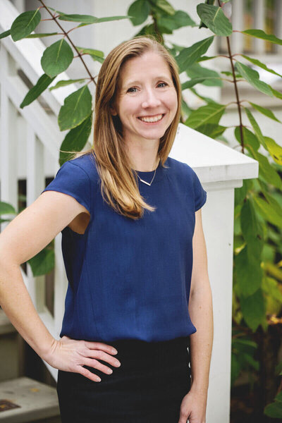 A woman named Lauren, wearing a blue shirt, stands confidently on a porch during daybreak, embodying a sense of wealth.