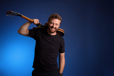 David Benfield, of First Coast News in his band,  in a black t-shirt holds an acoustic guitar over his shoulder against a gradient blue background, captured in the lively style of band photography Jacksonville.