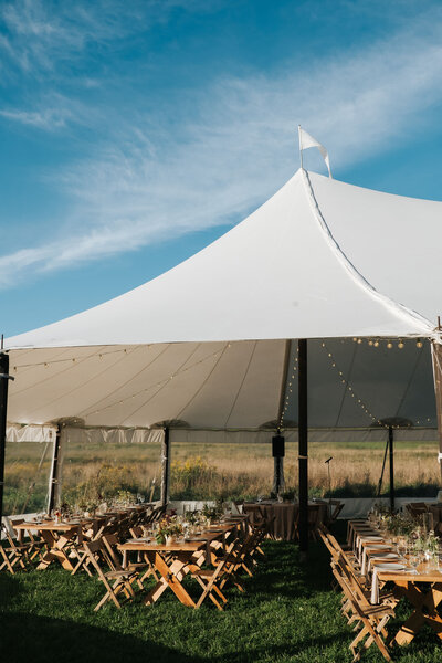 Blue skies over a sail-cloth tent  lined with twinkle lights with natural-wood tables and chairs on rich green grass.