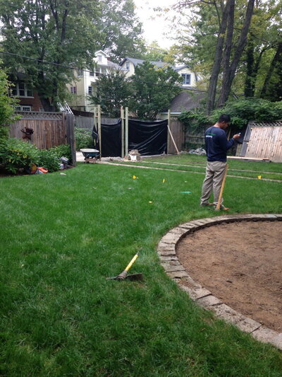 Backyard under construction with a partially dug-out circular area bordered by stones, workers preparing the space for a new patio installation.