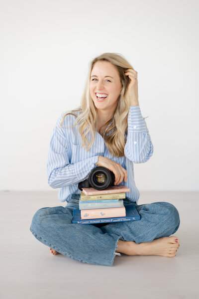 smiling woman tucks her hair behind her ear as she holds her photography camera and a stack of children's books while smiling at the camera