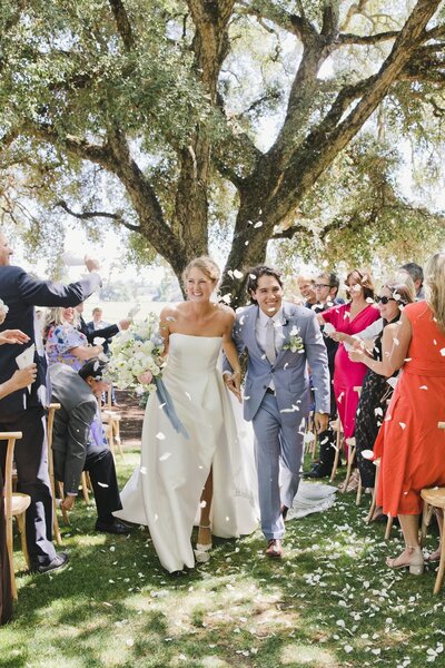 A fun wedding photo of a bride and groom walking down the aisle as their SoCal wedding guests shower them with flower petals.