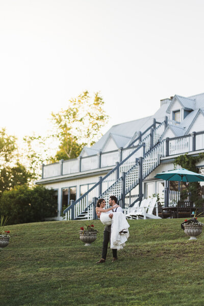 L’homme porte sa femme dans ses bras sous un magnifique coucher de soleil à l’Auberge Val Carroll, instant tendre et cinématographique capturé dans un décor naturel et romantique