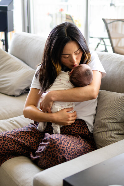 A tender moment of a mom cradling and kissing her newborn in cozy, natural window light – ellobelle photography.
