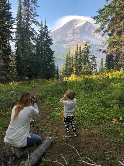 A woman and a child taking pictures of nature, with trees. The boy is wearing blue pants with white star print and gray t-shirt