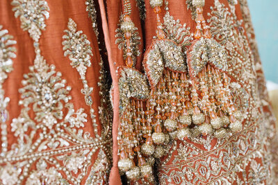 A stunning bridal lehenga in rich fabric, adorned with intricate gold hand embroidery that shimmers with every detail. Delicate tassels cascade gracefully, adding a touch of elegance and movement to the ensemble. The craftsmanship showcases traditional artistry, making it a breathtaking choice for a bride’s special day.