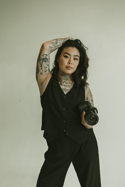 A tattooed woman wearing a black sleeveless outfit poses confidently while holding a camera, standing against a plain light background.
