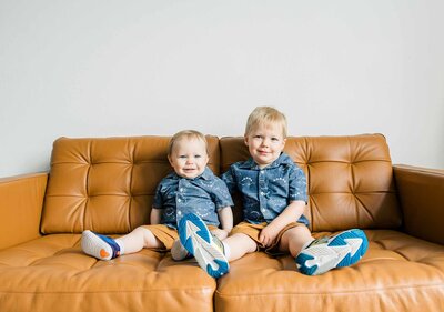 two little boys in matching shirts and shorts sit on a brown leather couch smiling