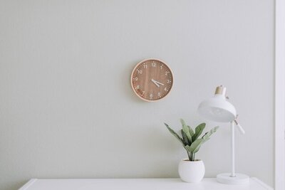 Image of a desk with a house plant and white lamp. There is a white wall background with a wood analog clock hanging on the wall.