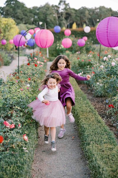 two little girls wearing pink dresses and running through a rose garden