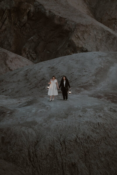 A couple walks hand in hand while hold lanterns to light their way through a dark path over the badlands