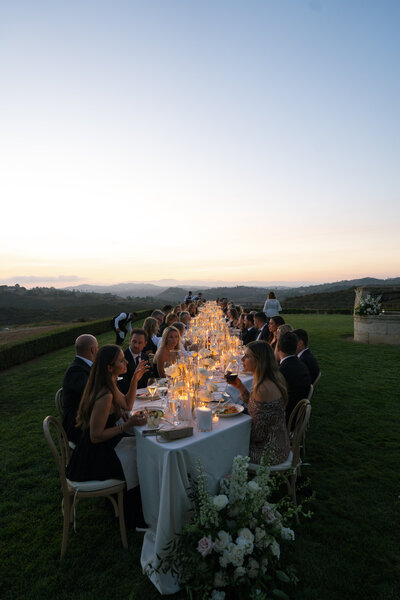 BP Collective European Inspired Experience with a long alfresco dining table set with white florals and candles against rolling hillside views.