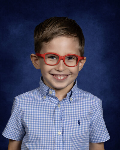 Smiling elementary school child wearing red glasses and a blue checkered shirt, posing against a solid blue background—cheerful and vibrant school portrait by Georgia school photographer Christina Barnett.