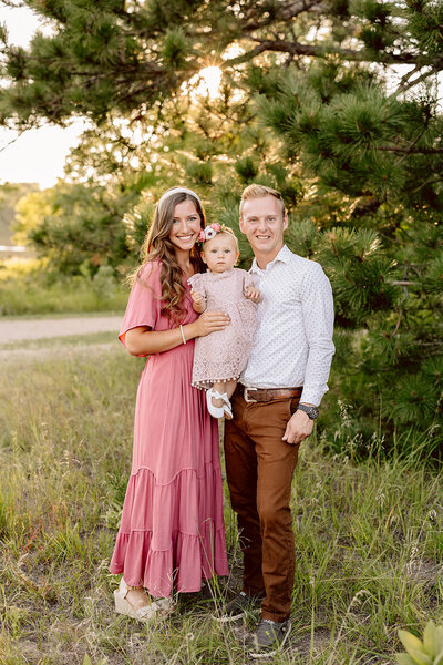 A family of three poses outdoors in a grassy area with trees, captured beautifully by a Brainerd Lakes Area Photographer. The woman, in a pink dress, holds their child in a matching outfit. The man wears a white shirt and brown pants. They smile as sunlight filters through the trees behind them.