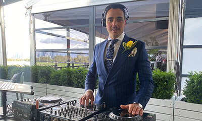Smiling DJ in a blue suit mixing music at an indoor event with branded DJ booth and lighting.