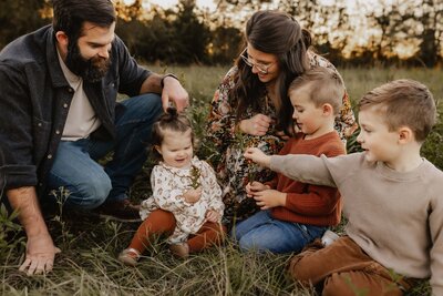 A family of five poses on the grass in a field, capturing the essence of Macon family photography. Two adults and three children joyfully interact, surrounded by lush greenery and trees in the background.