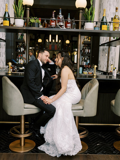 A couple in formal attire sits on barstools at a stylish bar, holding hands and gazing at each other. Shelves behind them display various bottles. Their enchanting moment is effortlessly orchestrated by a top-tier Canada wedding planner, adding an air of magic to the scene.