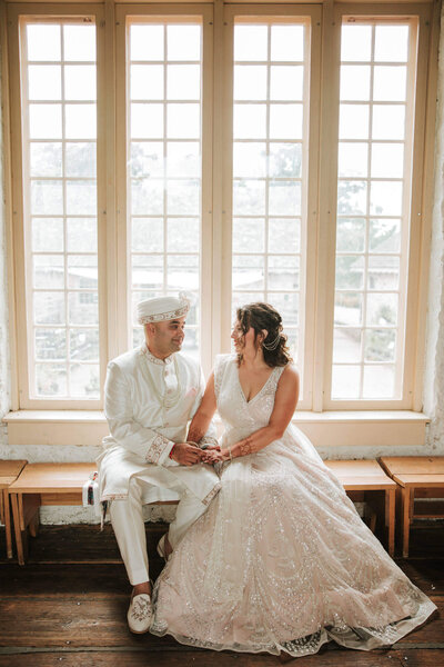 bride and groom sit together by a large window, holding hands and sharing a loving gaze in soft natural light.