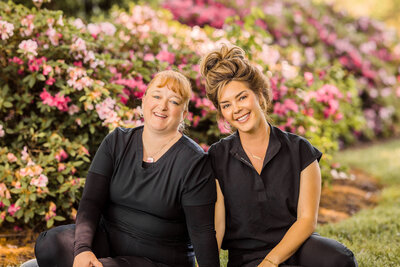 two women wearing black scrubs sitting in front of azalea bushes smiling