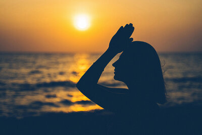 Woman in front of an orange sunset praying to the sky.