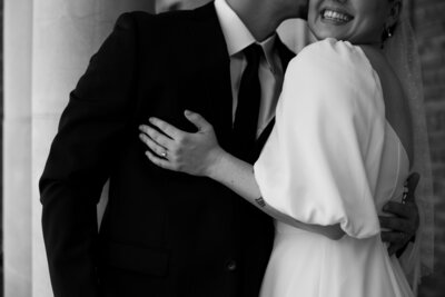 Close up crop bride and groom arms around each other, groom kissing on cheek