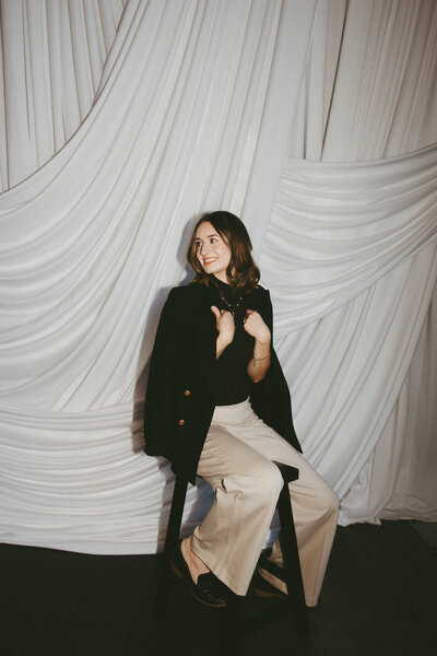 Customized calligraphy artist McKenna sits on a stool and looks over her shoulder as her hands grab her jacket collar.