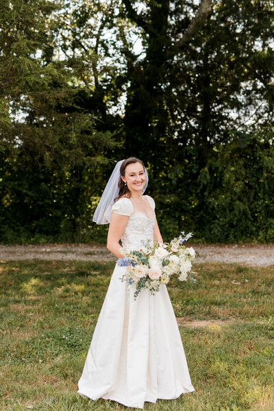 bride in a white lace gown and veil, smiling and holding a large bouquet, standing in a field