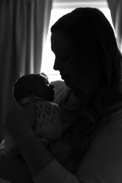 new mother with newborn daughter stand in front of window in shadow in black and white image