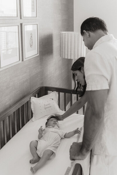 Black and white image of parents gently laying baby in crib, framed art in background.