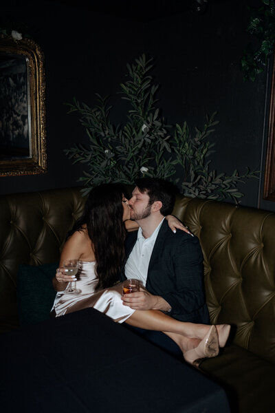 A couple cuddles up in a corner booth at a speakeasy.  Holding cocktails in their hand, they share a kiss during their engagement session.
