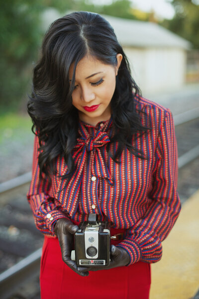 An Asian travel agent wearing a vintage outfit and standing by train tracks holding an antique Brownie camera for a personal branding photoshoot.
