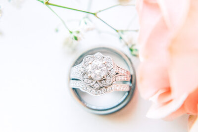 groom ring laying down on a flat white surface with brides ring propped up showing the stones in the engagement ring surrounded by flowers