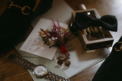 Éléments de décor de mariage comprenant une montre, un faire-part, un parfum, un noeud papillon, des fleurs séchées.