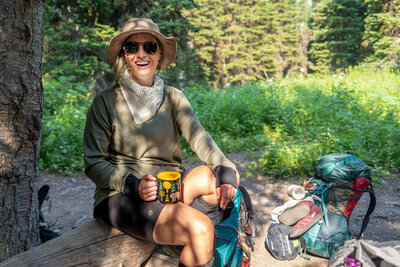 A woman wear Smartwool shorts and a Smartwool bucket hat sits drinking coffee at her campsite in Glacier National Park, Montana.
