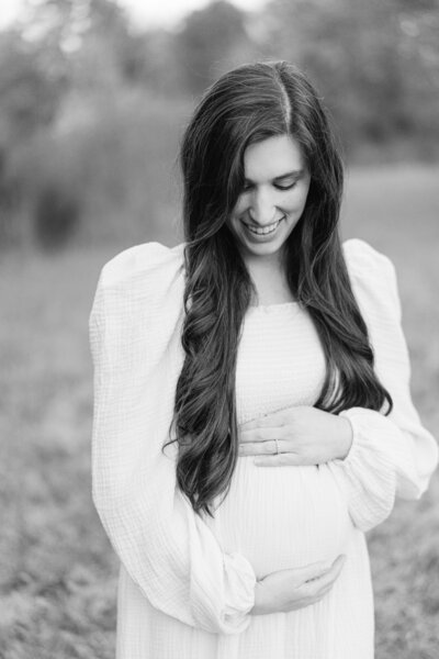 A pregnant woman is wearing a white maternity dress and laughing at the camera for her maternity photos with nashville family photographer Dolly DeLong Photography
