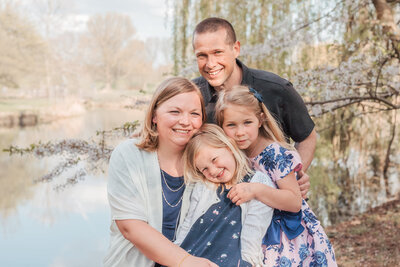 Family of four. Mom, dad and two girls.  Background: a weeping willow, cherry trees in bloom and a body of water. Youngest girl snuggling into mommy. Everybody close. Dad hugging his laddies from behind. Family Photography - cherry blossom session