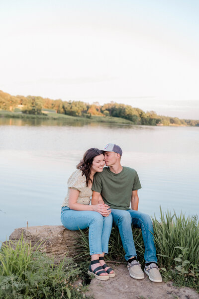 Couple poses for the camera on a rock near the lake with the man kissing the woman's cheek.