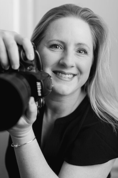 Black and white headshot of Kristin Palkoner holding her Nikon camera and smiling.