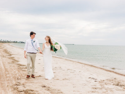 A bride and groom walk hand in hand along a sandy beach, with the bride holding a bouquet and her veil blowing in the breeze. The ocean and cloudy sky are in the background.The couple is a wedding photography team in Austin.