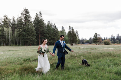 A groom. wearing a blue suit, has the leash to his chocolate lab in one hand while holding the hand of his bride . She is wearing a white gauzy gown and holding a big bouquet of flowers as they stroll through the tall green grass at their Black Butte Ranch elopement in Oregon. | Erica Swantek Photography